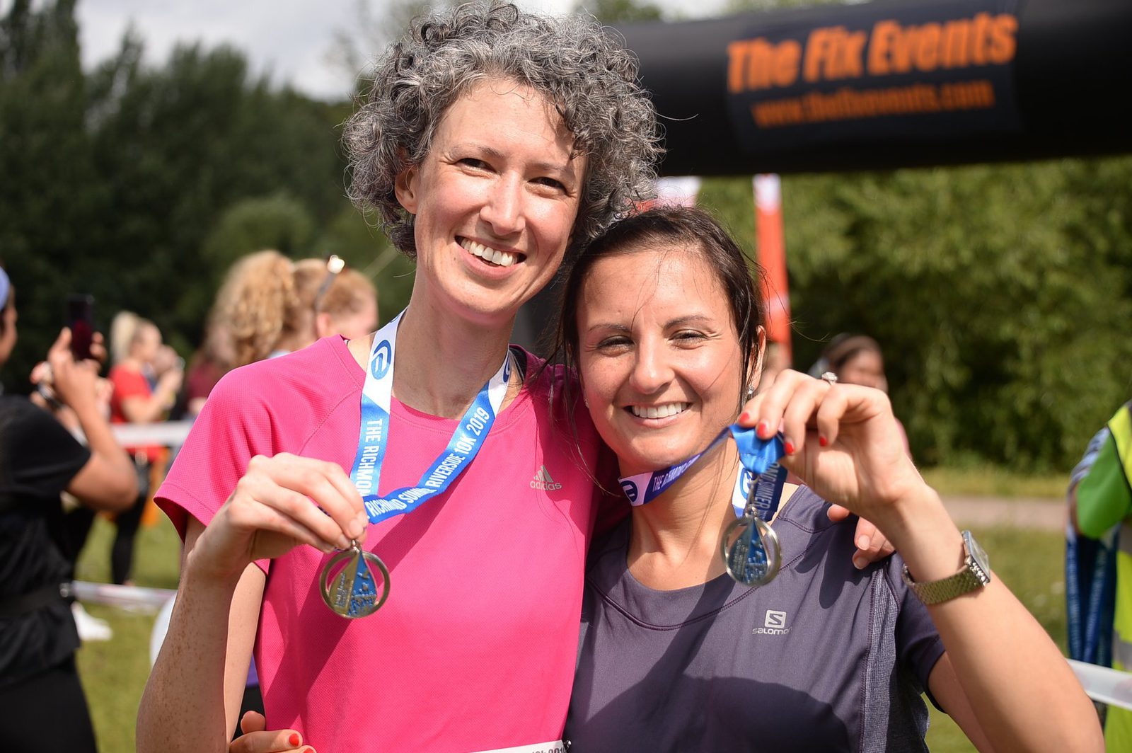 Two smiling women hold up medals after a race. The woman on the left has short curly gray hair and is wearing a pink shirt, while the woman on the right has dark hair and a gray shirt. They are standing outdoors with other people and a black event banner in the background.