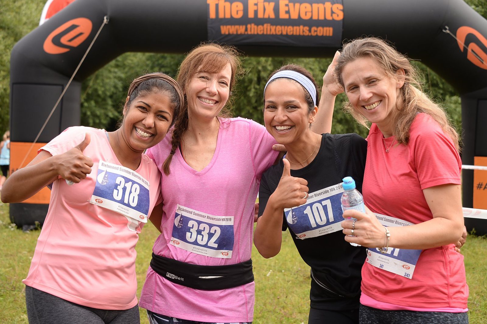 Four women standing together and smiling after a race, wearing race bibs 318, 332, 107, and 119. One holds a water bottle, and another gives a thumbs-up. They are in front of a black and orange inflatable arch that reads "The Fix Events.