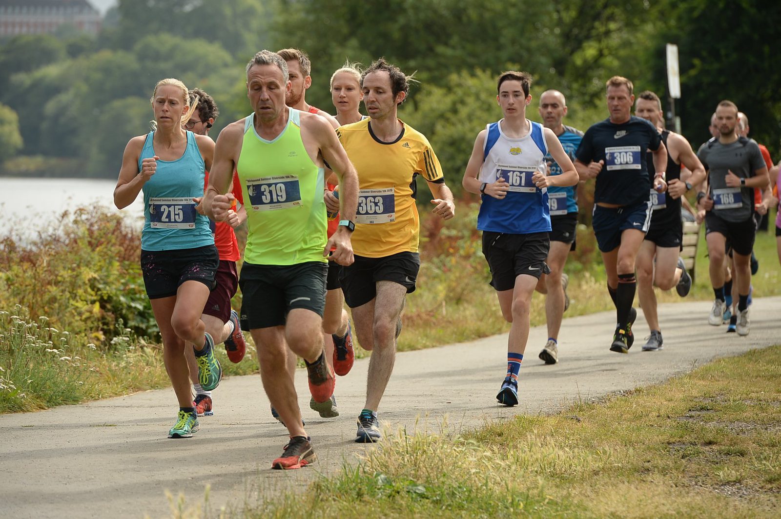 A group of runners, wearing bib numbers, race along a paved path by a lake. The participants are dressed in athletic gear of various colors, and some wear shorts and tank tops. Trees and greenery line the path, with a hint of a building in the background.