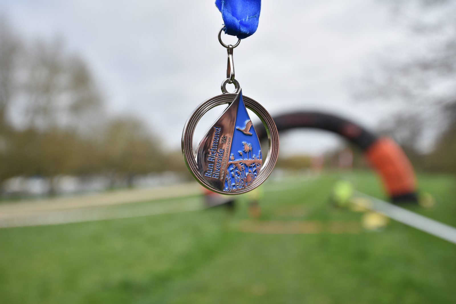 A blue and silver medal hangs from a blue ribbon in the foreground, with the words "Richmond Marathon" and an intricate design that includes an athlete running. The background features a blurred grassy area and an arch, indicating a finish line.