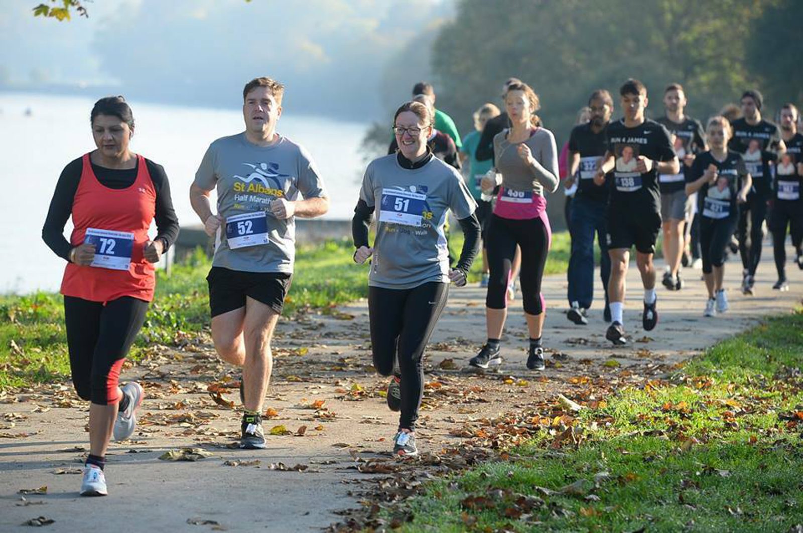 Runners participate in a race on a paved path beside a body of water on a sunny day. Fall leaves are scattered on the ground. The participants, wearing numbered bibs, appear focused and determined as they run. Trees with green foliage line the pathway.