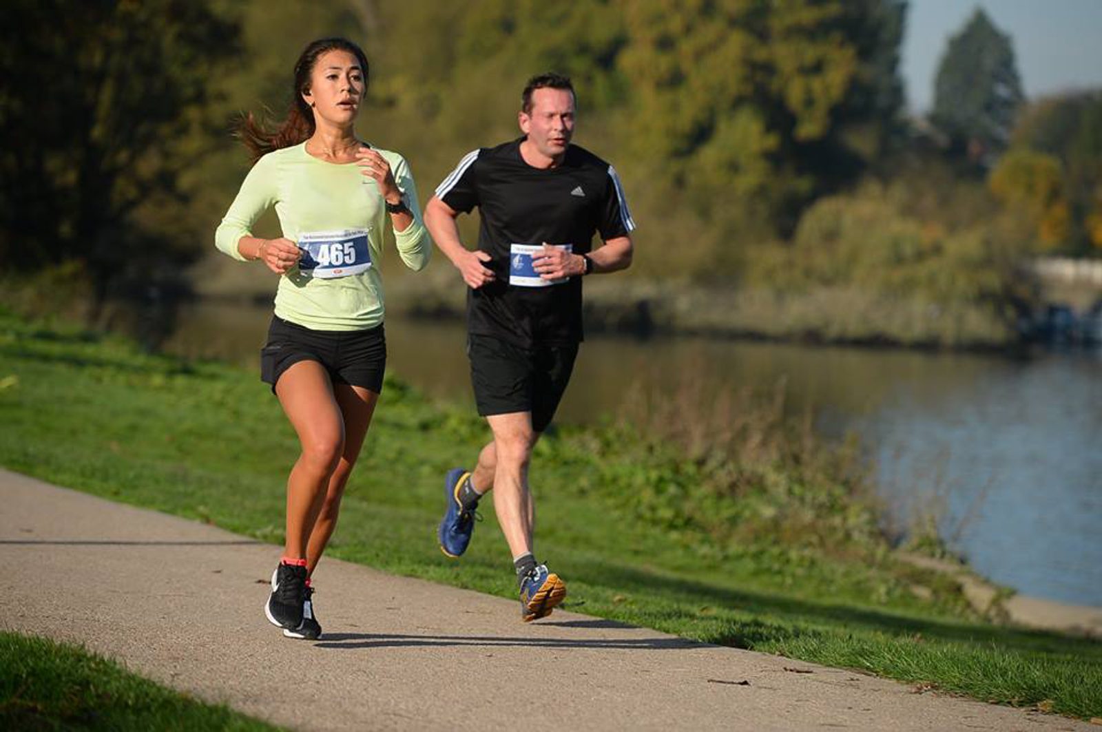 Two runners are jogging on a paved path beside a body of water in a park. The woman in front is wearing a light green top and black shorts, and the man behind her is wearing a black shirt and shorts. Both have race bibs and are focused on their run. The background is green and lush.