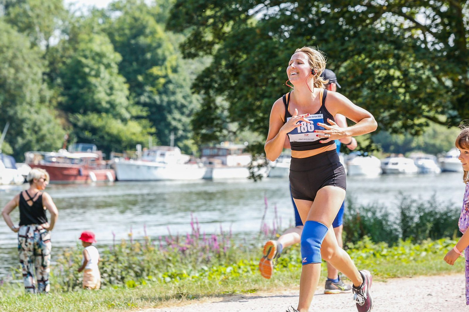 A woman wearing a black sports bra and shorts, with a blue knee brace, is running in a race by a riverside. She is smiling and wearing earphones. Other people can be seen in the background, including children and boats on the river.