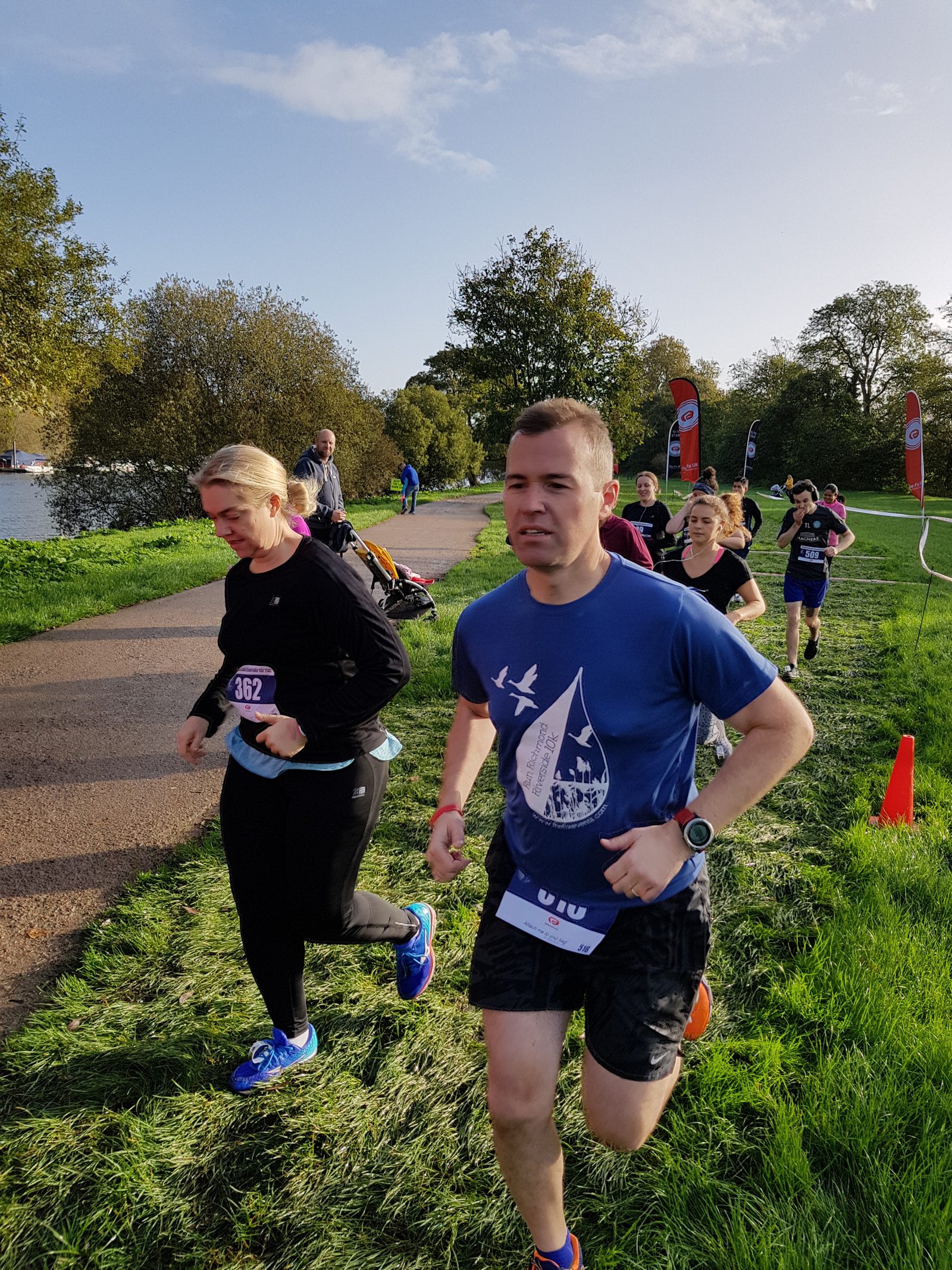 A group of runners is participating in a race along a scenic, tree-lined path near a body of water. The sunny weather casts shadows on the grassy area beside the path. Orange flags and a few spectators can be seen in the background.