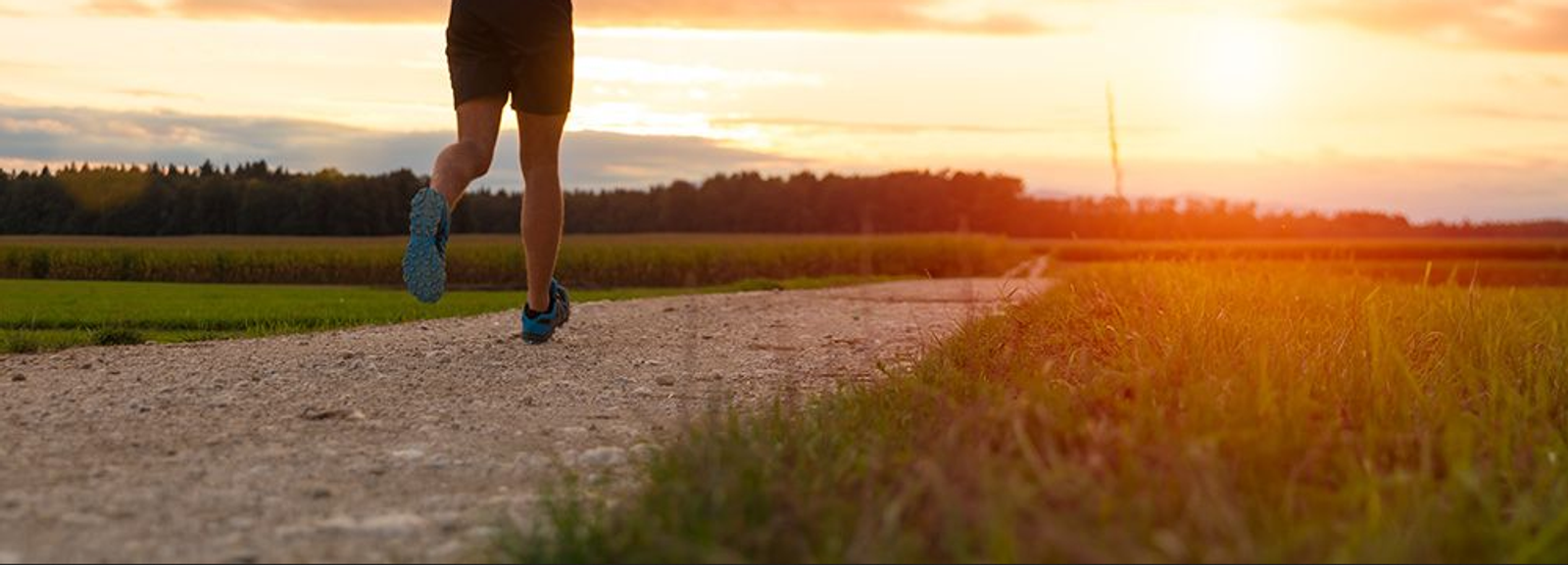 A person running along a gravel path during sunset in a rural area. The sun is low in the sky, casting a warm, golden light. The path is flanked by grassy fields and trees in the background. The runner wears shorts and sneakers.