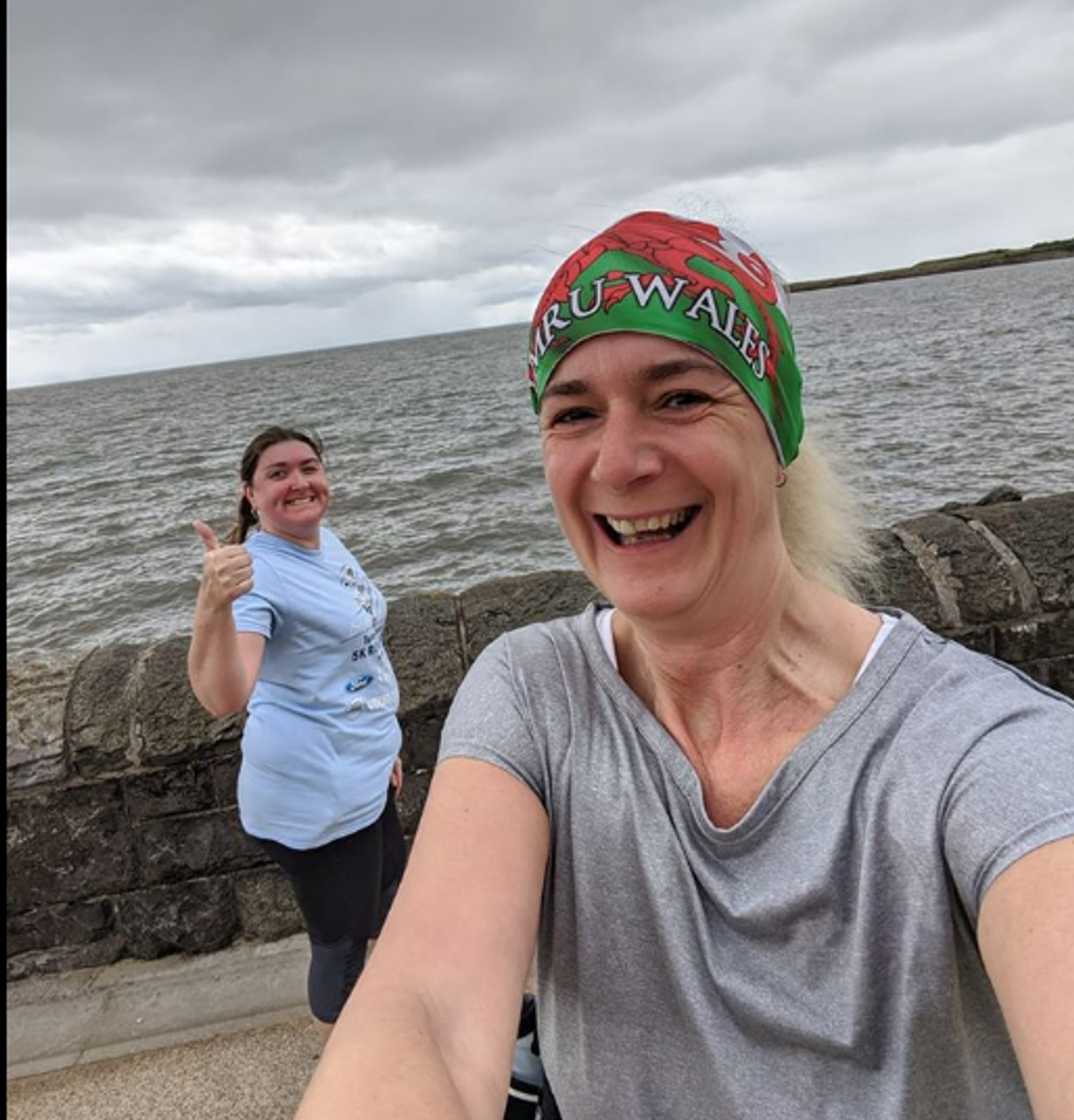 Two women are smiling and posing for a photo by the seaside on a cloudy day. The woman in front is wearing a gray shirt and a red and green headband that reads "WALES," while the other woman is giving a thumbs-up and wearing a light blue shirt. The sea and a stone wall are visible in the background.