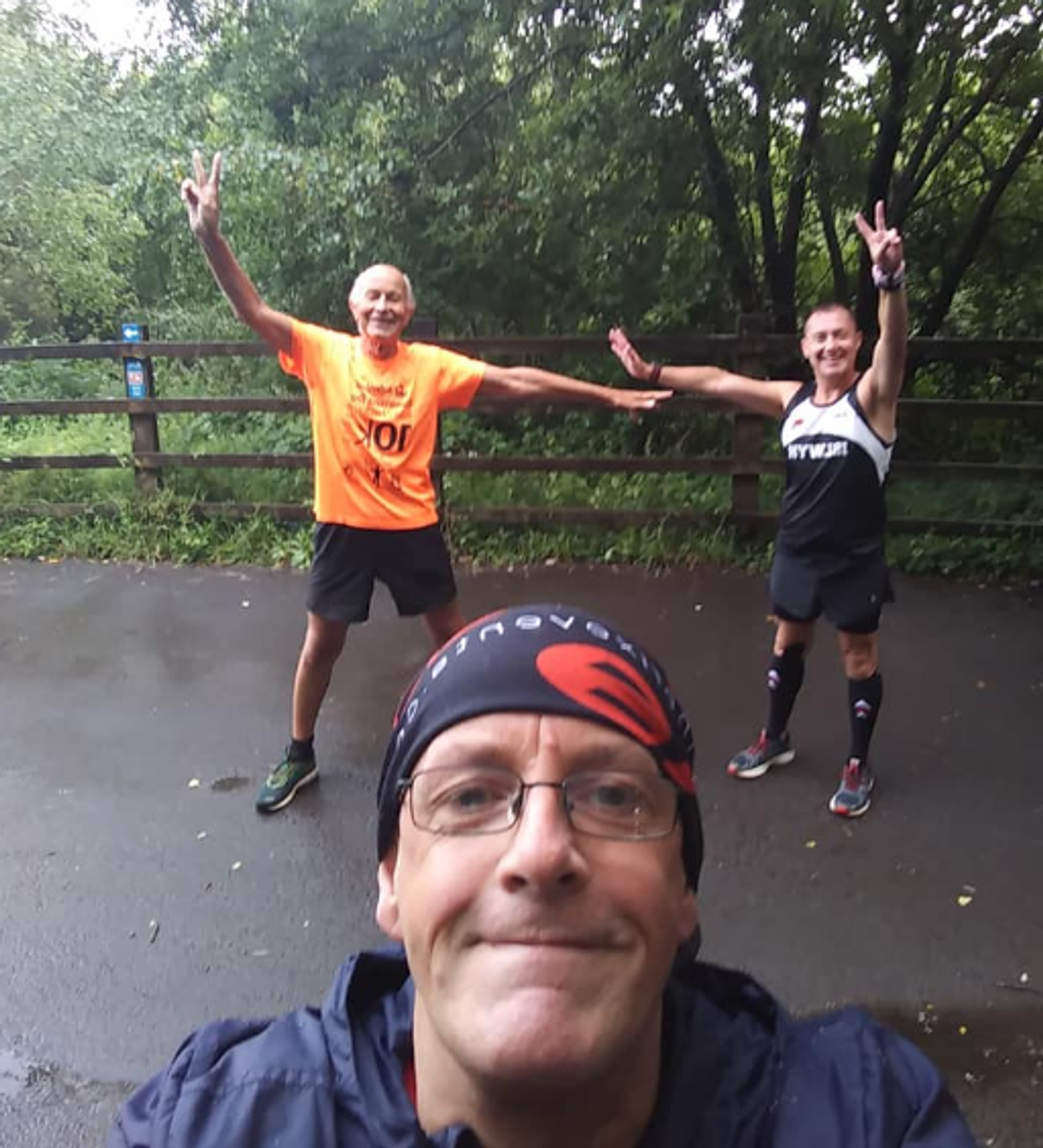 Three people are posing for a photo outdoors. The person in the foreground is wearing a hat and glasses, taking a selfie. Two people in the background are smiling with their arms outstretched and flashing peace signs. Trees and a wooden fence are visible in the background.