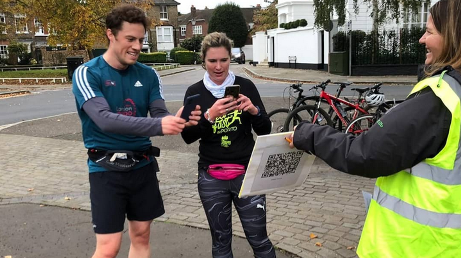 Two runners, a man and a woman, pause to scan a QR code held by a volunteer in a reflective vest. They are on a paved path with bicycles parked nearby and houses in the background. The man is smiling and the woman is focused on her phone.