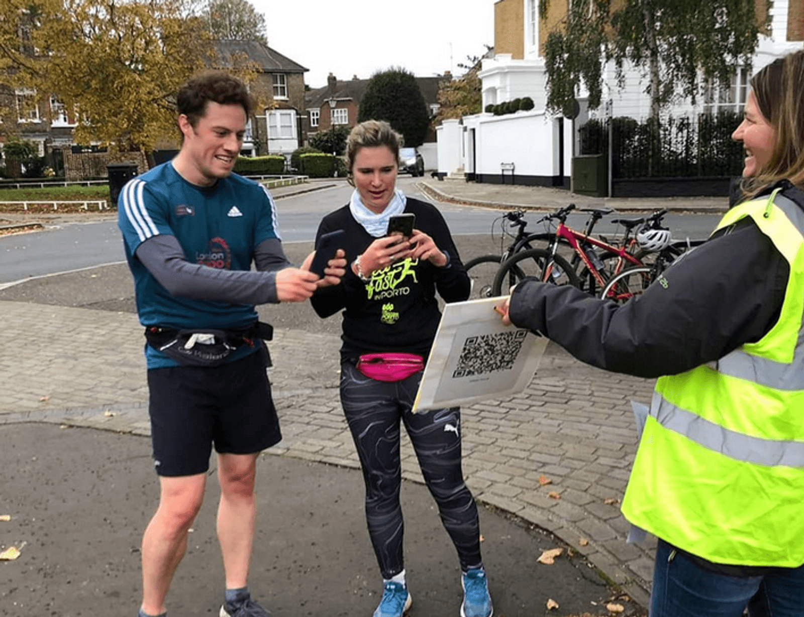 Two people in athletic wear are shown outside, engaging with a woman in a yellow safety vest. The man appears joyous, gesturing toward a signboard with a large QR code that the woman in athletic wear is scanning with her phone. Bicycles and houses are in the background.