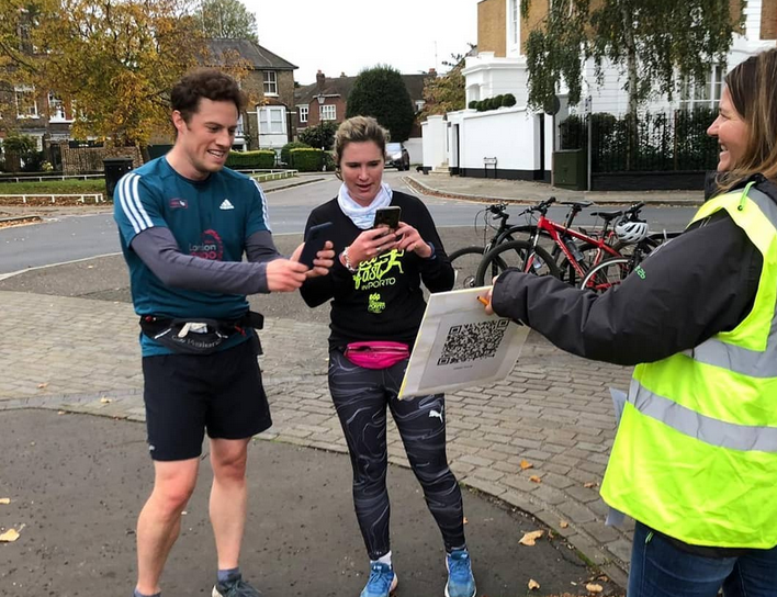 Two people in athletic wear are shown outside, engaging with a woman in a yellow safety vest. The man appears joyous, gesturing toward a signboard with a large QR code that the woman in athletic wear is scanning with her phone. Bicycles and houses are in the background.