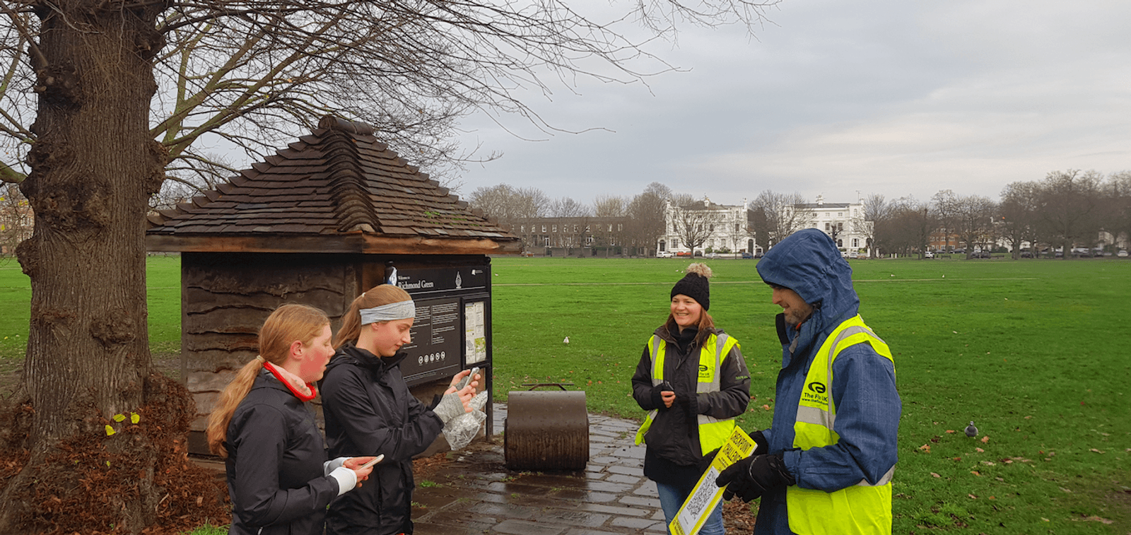 Four people in a park are participating in an outdoor activity. Two wear yellow high-visibility vests with one holding a clipboard; another holds a stopwatch. Two others, dressed for cold weather, appear to be engaging with the organizers near a large tree and a small wooden structure.