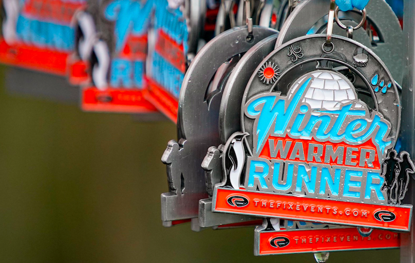 Close-up view of multiple Winter Warmer Runner medals hanging. The medals feature an igloo and snowflakes, with large blue and orange "Winter Warmer Runner" text, and thefixevents.com written on them.