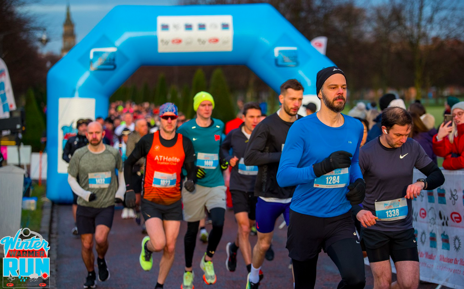 A group of runners participating in a winter marathon is captured mid-race. They are wearing athletic gear, some with long sleeves and others with shorts, running towards the finish line marked by a large blue inflatable arch. The weather appears cold and the race logo is visible.