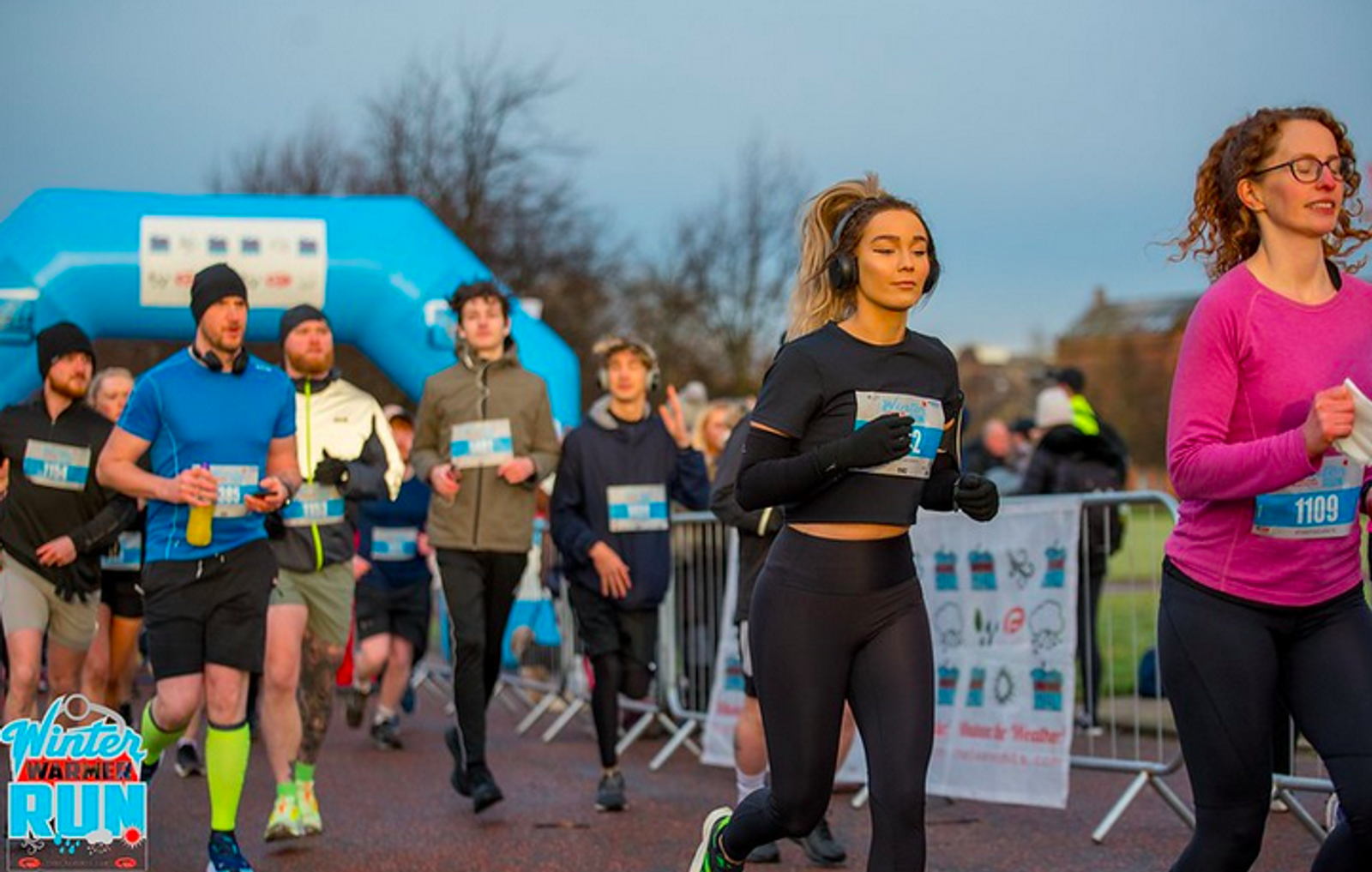 A group of runners participates in the Winter Run event. They are running on a paved path with some bundled up in warm clothing, while others wear athletic gear. An inflatable blue arch and barricades line the route. The mood is energetic and focused.