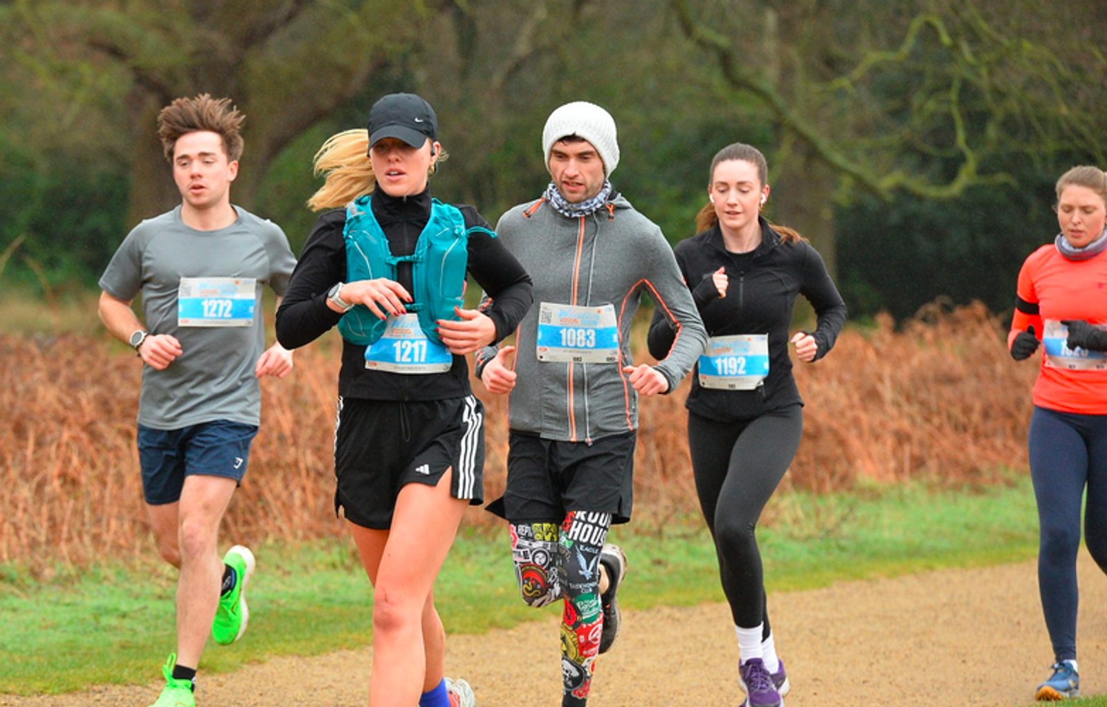 A group of runners participating in a race on a dirt path in a park. They wear athletic clothing and numbered bibs. Trees and dry grass are visible in the background, and the weather appears overcast.