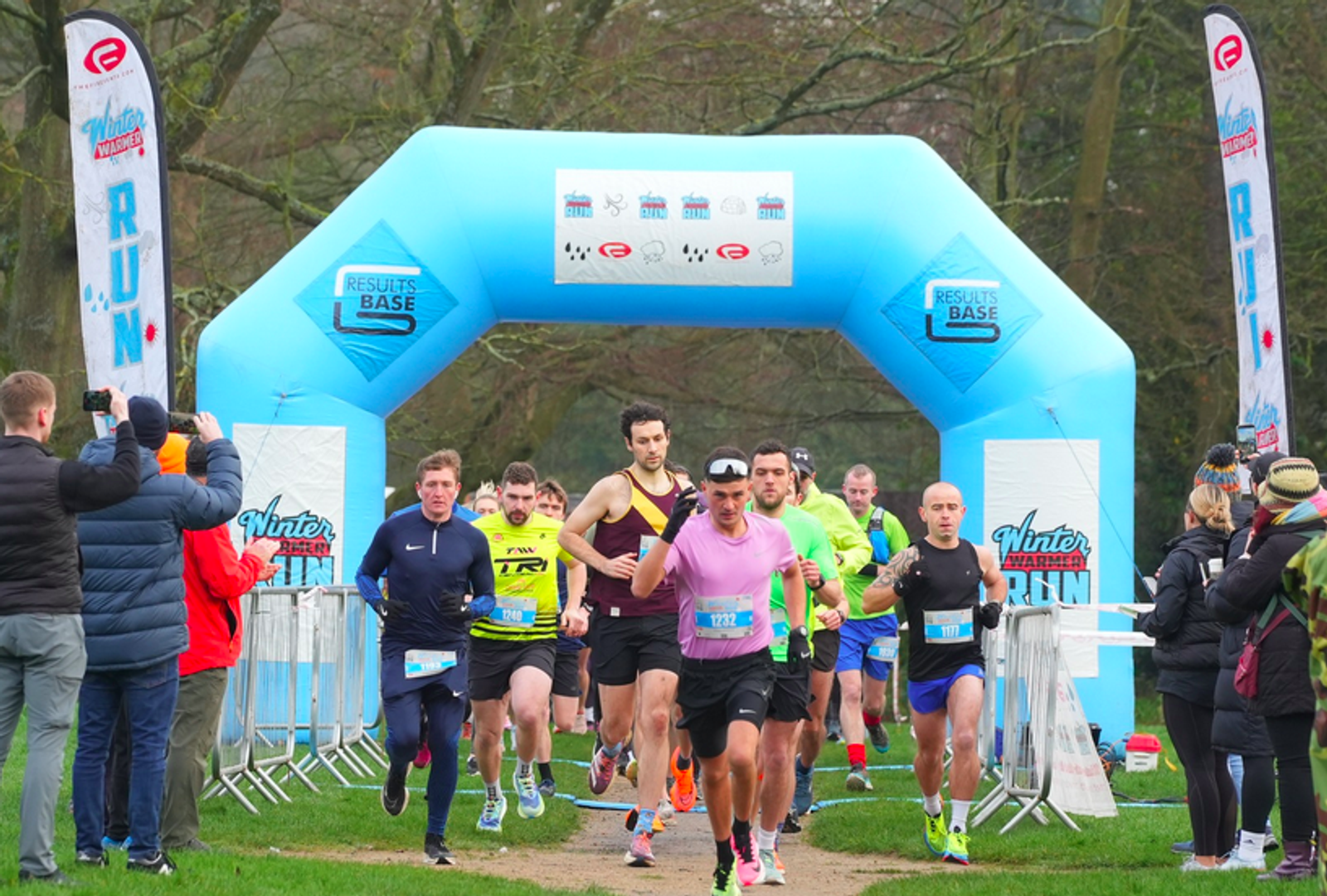 Runners in colorful athletic wear start a race, passing under a blue inflatable arch with "Winter Run" branding. Spectators and photographers line the path, capturing the event on a cloudy day in a park setting.