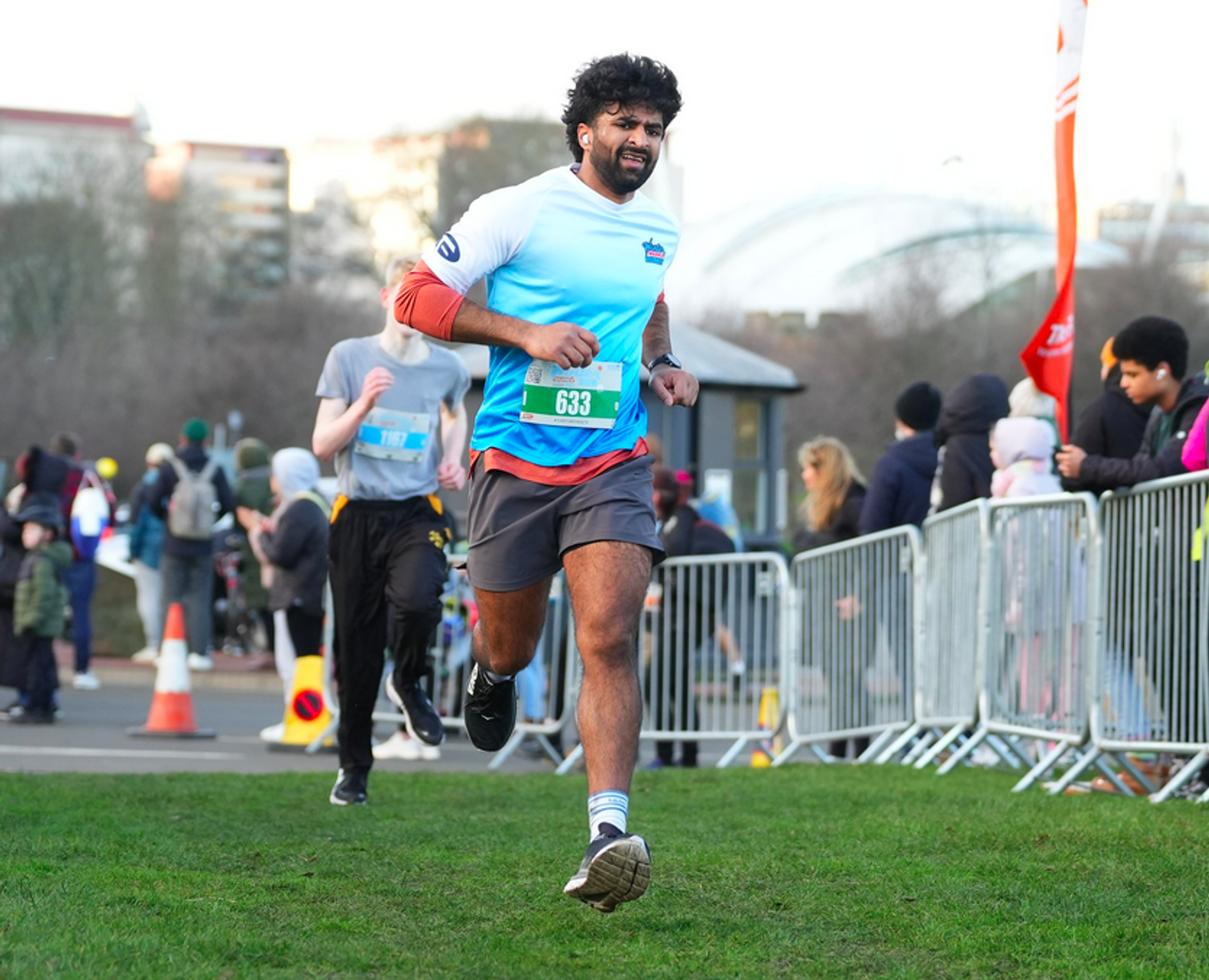 A runner wearing a light blue shirt and gray shorts sprints towards the finish line in a park race. He's competing in a diverse crowd, with spectators behind barriers cheering. The sky is clear, and city buildings are visible in the background.