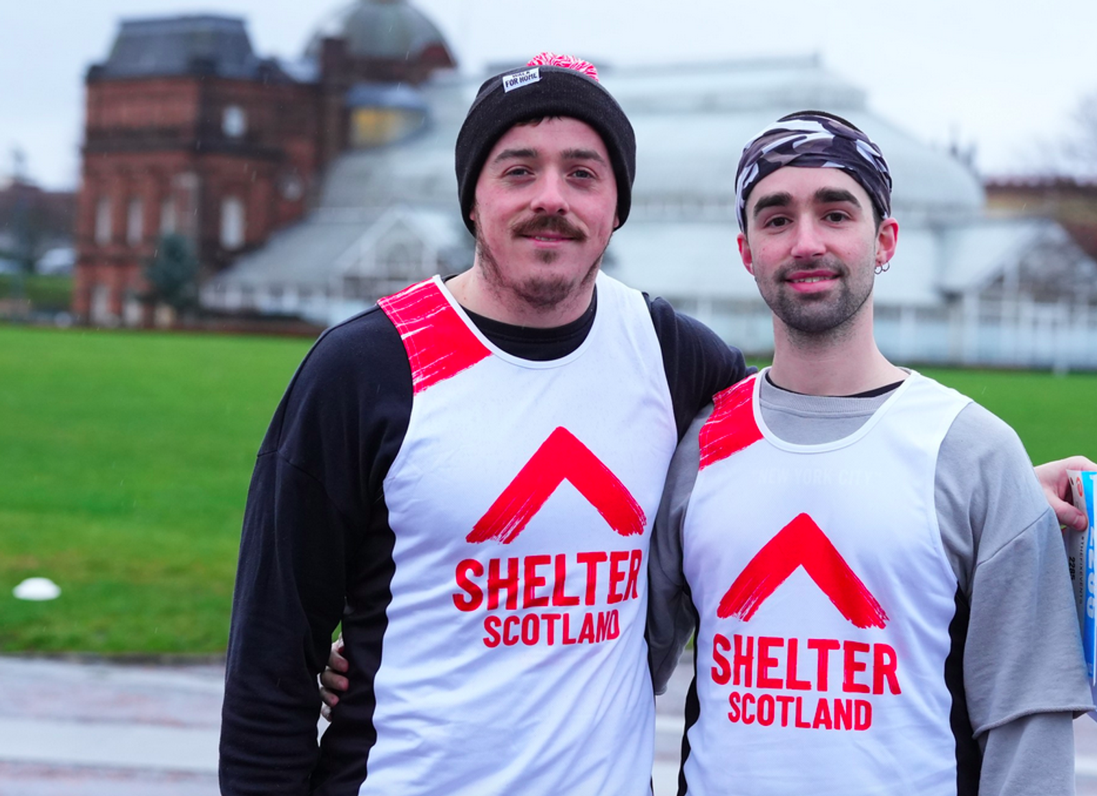 Two people standing outdoors wearing matching white vests with red and black graphics that read "Shelter Scotland." They are in front of a grassy area and a large building. One wears a black beanie, and the other has a patterned headband.