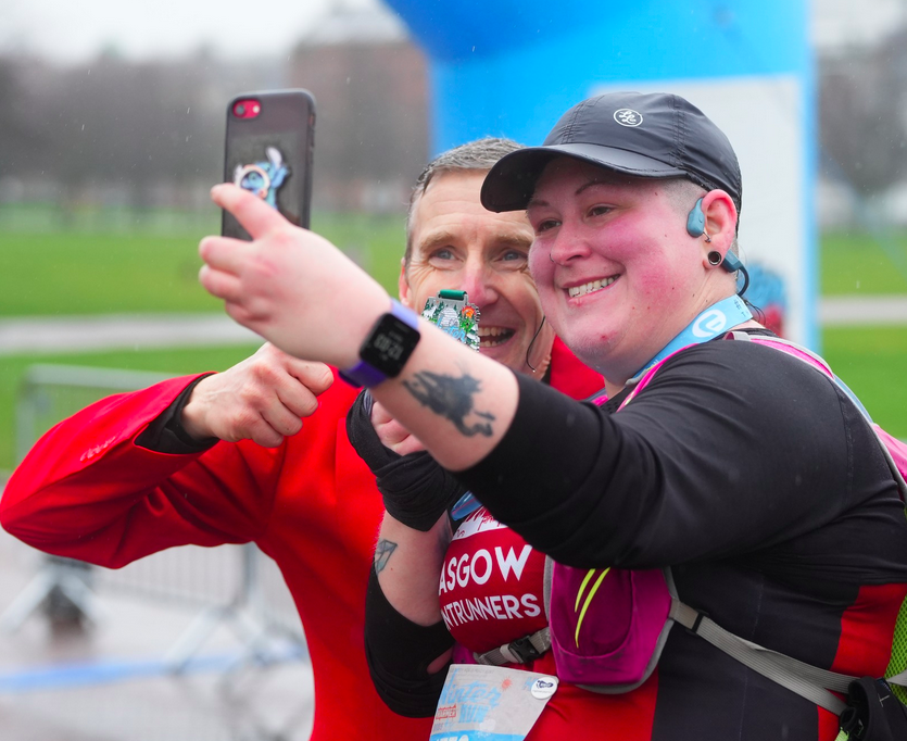 Two smiling runners take a selfie at the finish line of a race. One holds up a medal, and they both seem happy and celebratory. The background shows an outdoor event setting.