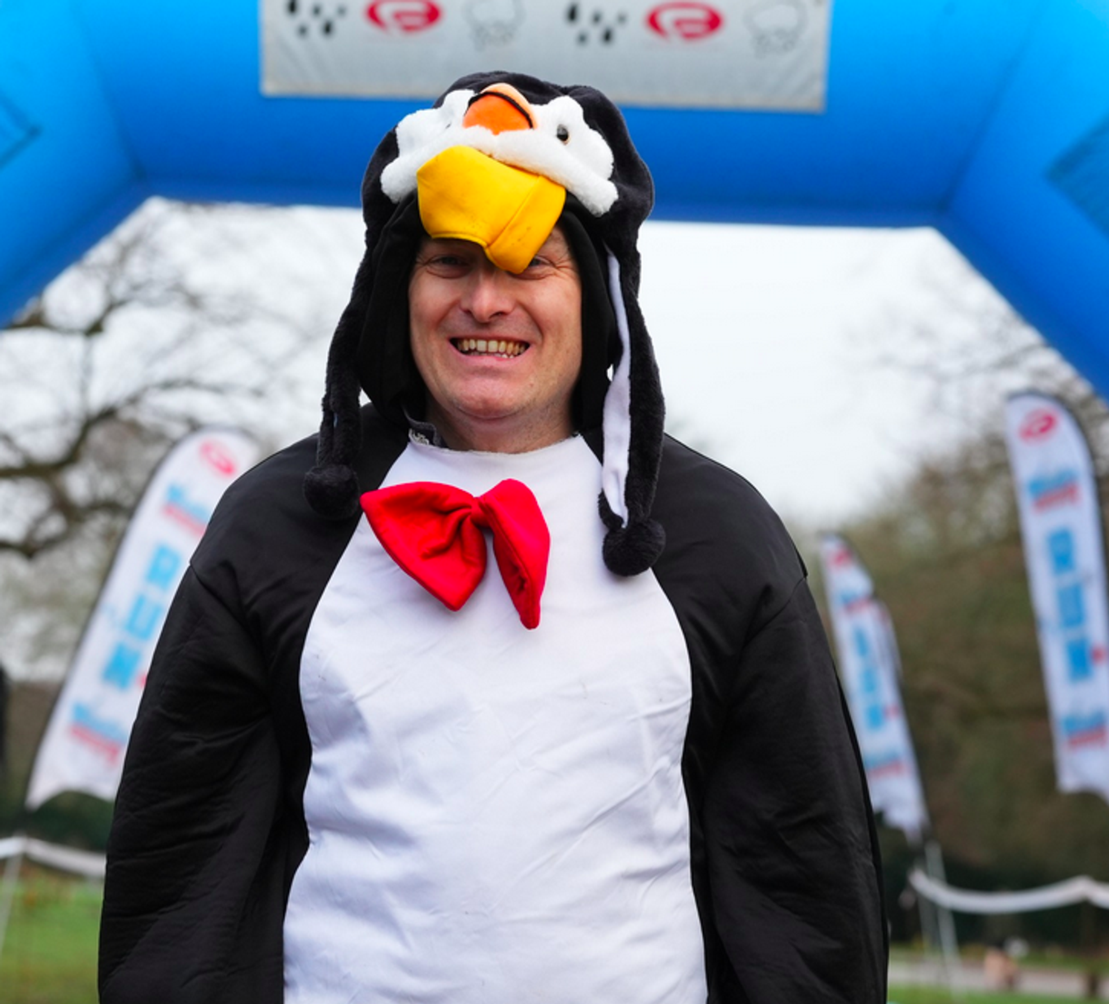 Person in a penguin costume with a red bow tie smiling under a blue inflatable archway. White banners with red and blue symbols are in the background.