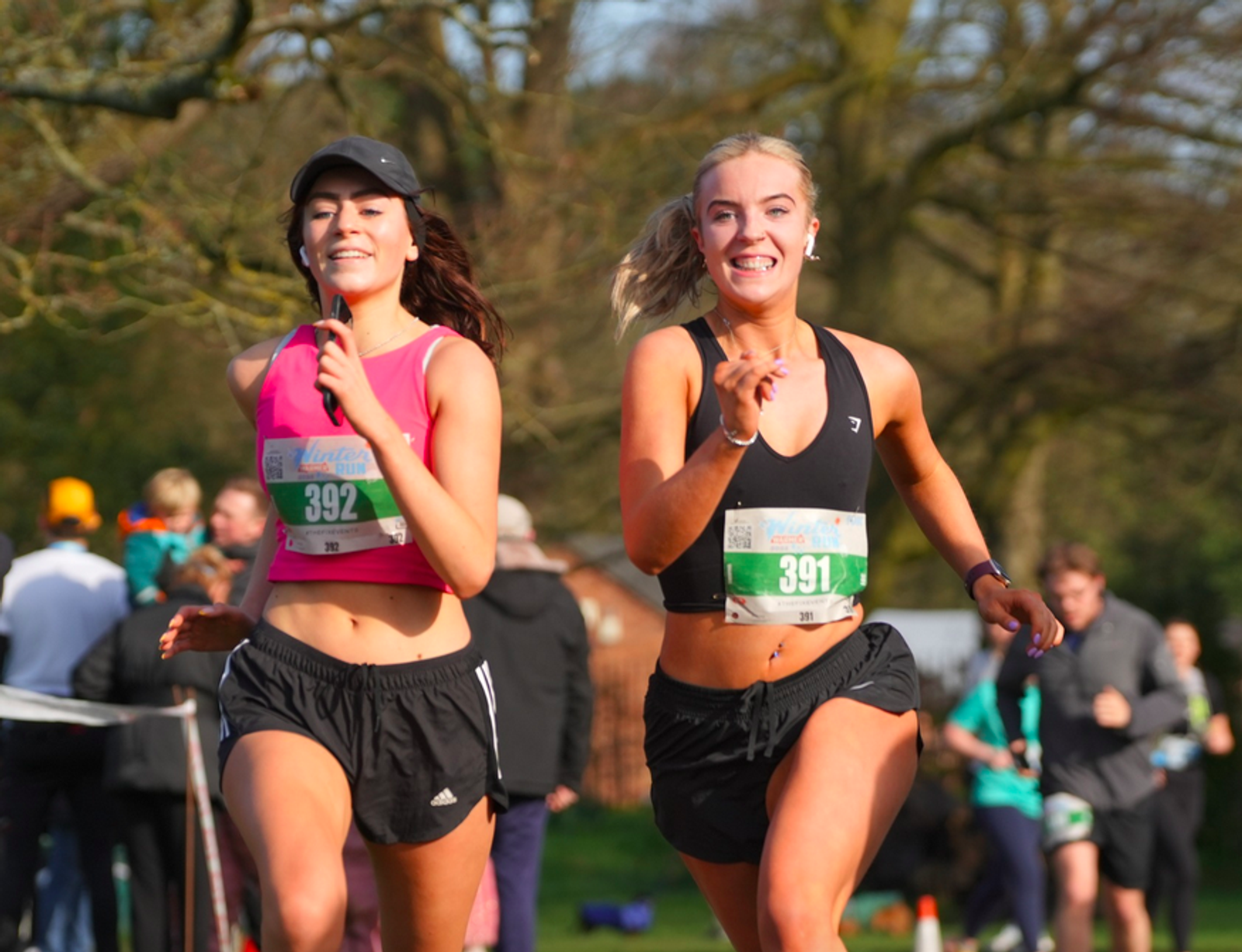 Two women are running in a race, smiling and wearing athletic gear with numbered bibs. They are outdoors with fellow runners and trees visible in the background. It's a sunny day.
