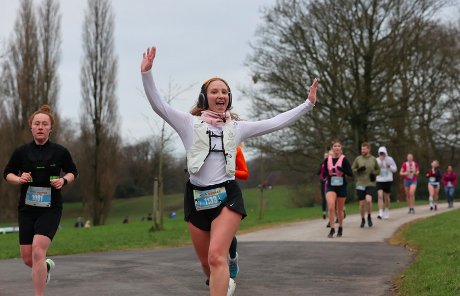 A woman joyfully runs towards the camera with arms raised, wearing a race bib. Other participants are seen running behind her on a pathway, surrounded by grass and leafless trees. The sky is overcast.