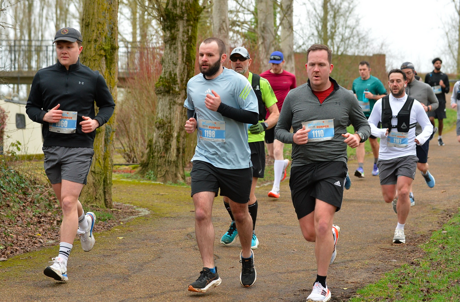 A group of runners participating in a race on a tree-lined path. They are wearing numbered bibs. The weather appears cool, with some participants wearing long sleeves and caps. The background shows trees and a few spectators.