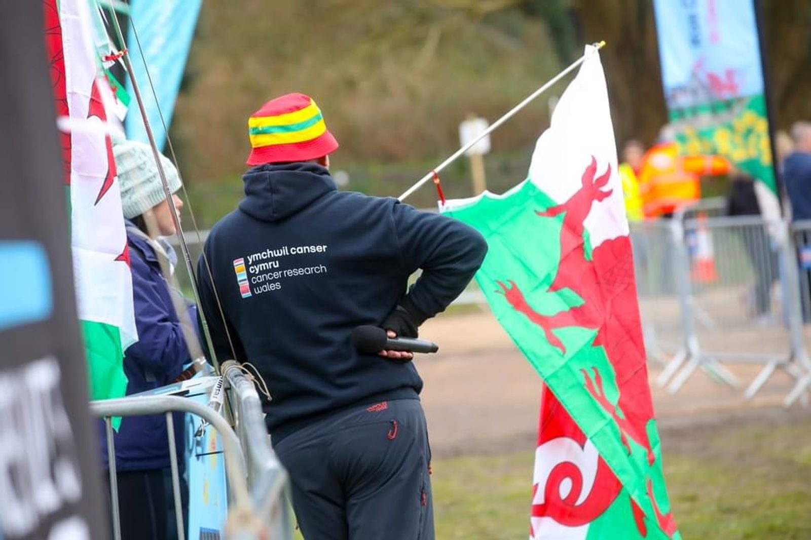 A person wearing a colorful striped hat and a dark hoodie with cancer-related text stands holding a large Welsh flag. They are turned away from the camera, observing an outdoor event with barriers and attendees in the background.