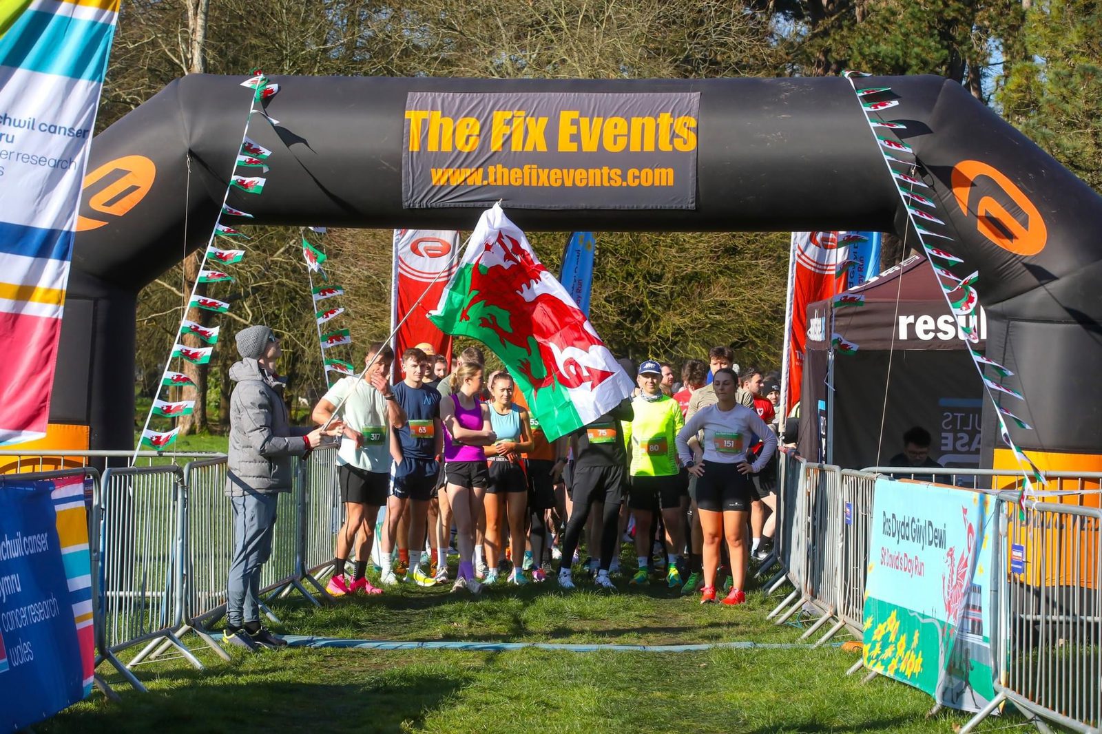 Runners gather at the start line under an inflatable archway reading "The Fix Events." The group stands behind a person holding a large flag with a red dragon. Colorful banners and a sunny, green outdoor setting complete the scene.