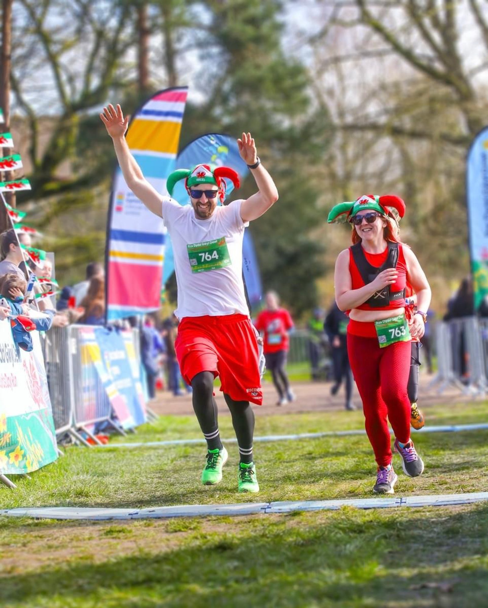 Two runners dressed in festive outfits, including colorful hats, are cheerfully crossing a finish line in a race. The man in white shirt and red shorts raises his arms in victory, while the woman in red attire and sunglasses smiles beside him. Trees and banners are in the background.