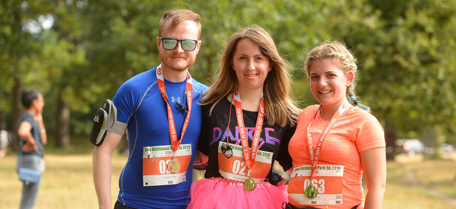 Three runners pose for a photo after a race in a park. They are wearing race bibs and medals around their necks. The runner on the left is dressed in a blue outfit, the middle runner in a black top and pink tutu, and the runner on the right in an orange top. Trees are in the background.