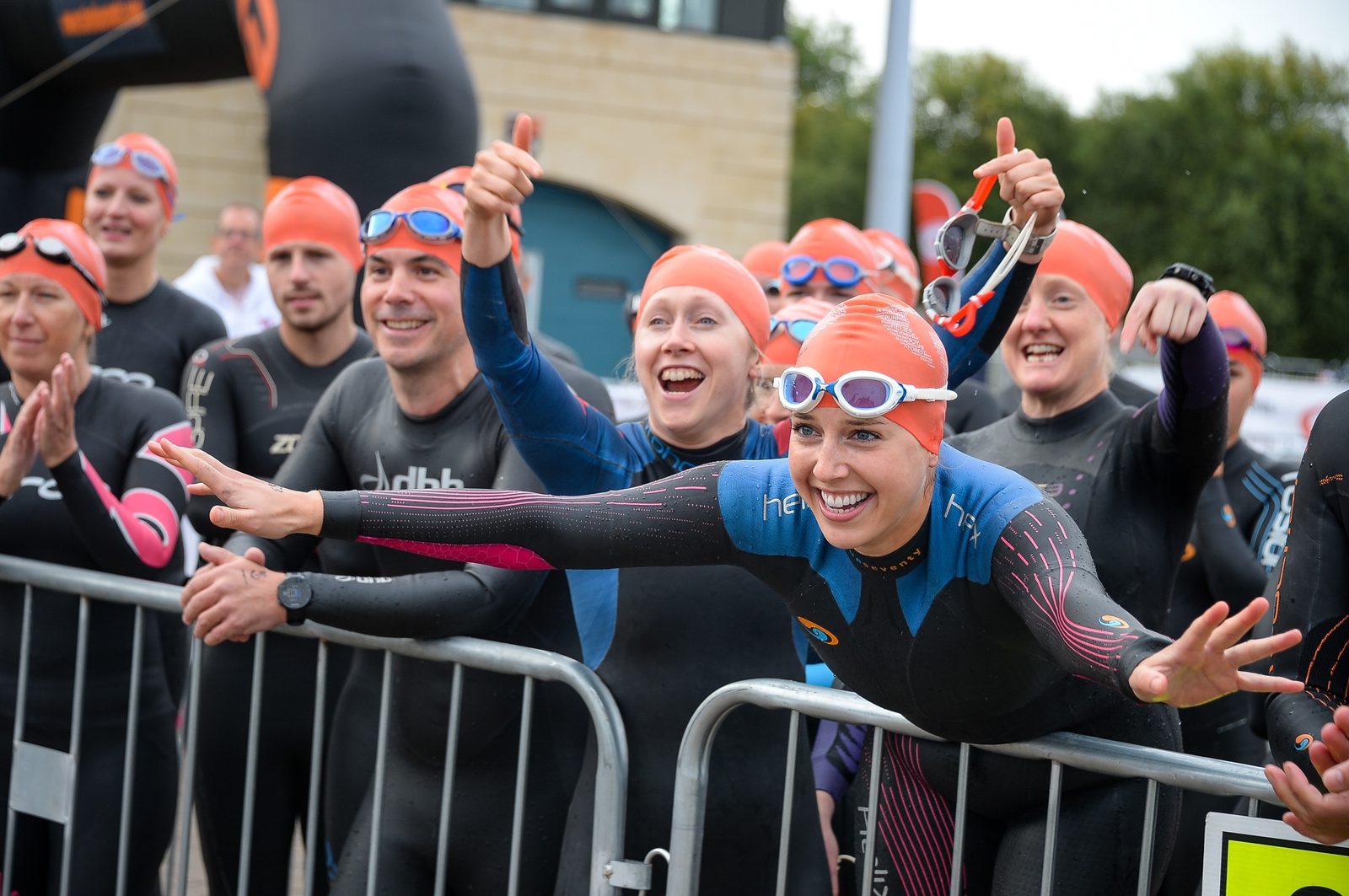 A group of enthusiastic triathletes in wetsuits and orange swim caps stand behind a metal barrier, cheering and smiling before a race. One woman in front, wearing goggles, energetically reaches forward with excitement.