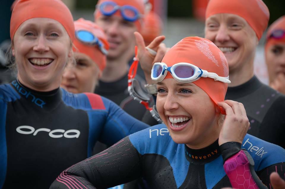 A group of swimmers wearing wetsuits and orange swim caps, preparing for a swim. They are smiling and adjusting their goggles and caps, looking happy and excited.