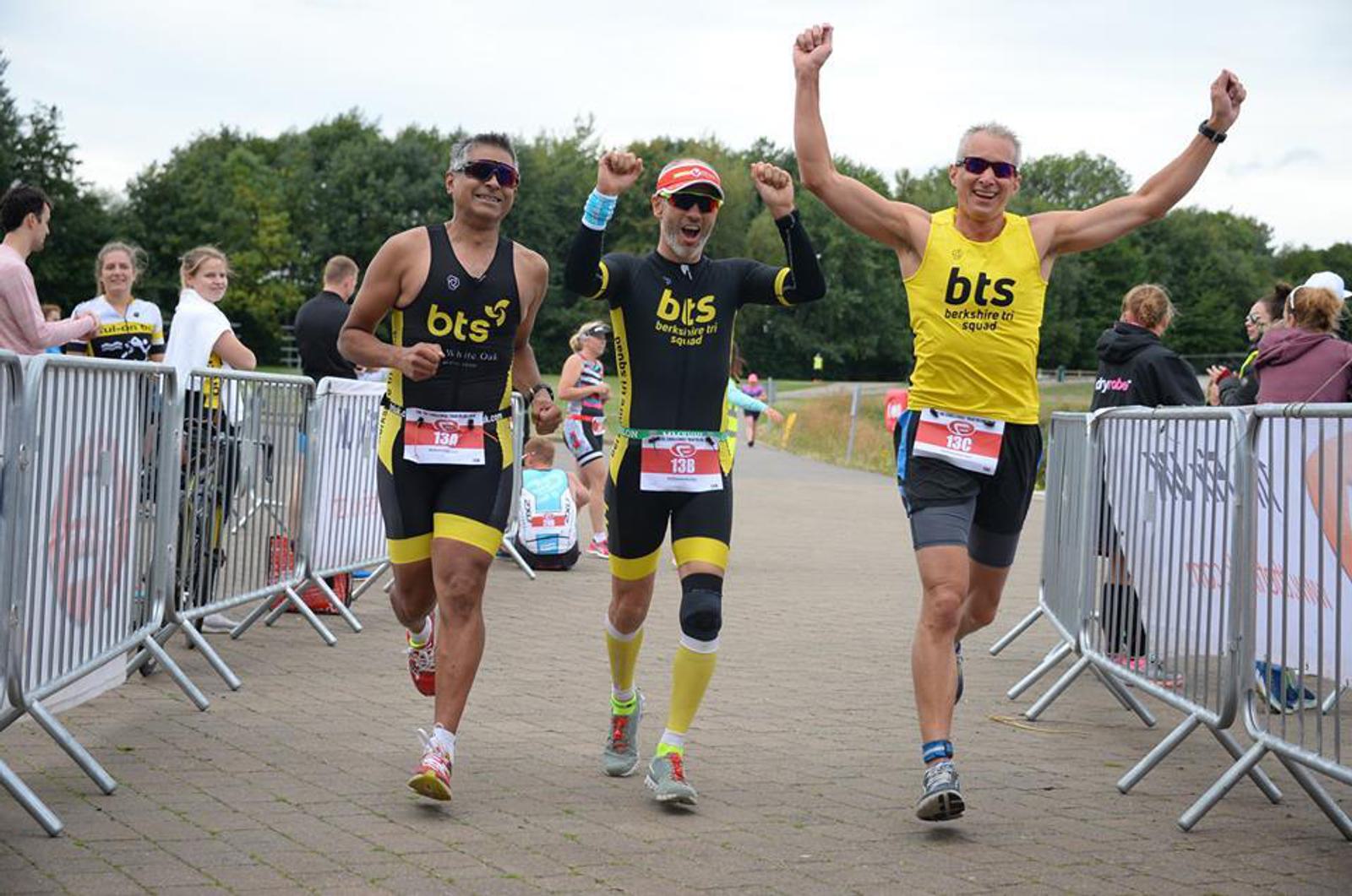 Three athletes in matching black and yellow outfits celebrate as they cross the finish line of a race. They are smiling and raising their arms triumphantly. Onlookers and barriers line the path on a cloudy day.