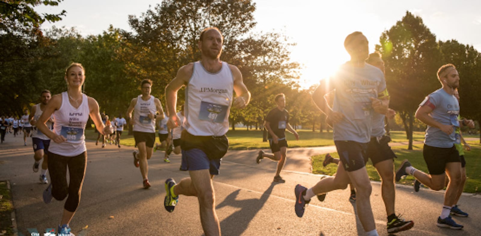 A group of runners participating in an outdoor race on a sunlit path surrounded by trees. The sun is low in the sky, casting a warm glow over the runners. Some are dressed in white tank tops and shorts, while others wear various athletic attire.