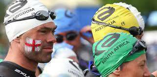 Several swimmers wearing swim caps and goggles prepare for a race. The swimmer in the foreground has an England flag painted on his face and is wearing a white cap that says "KINGDOM." Caps for Australia and South Africa are visible in the background.