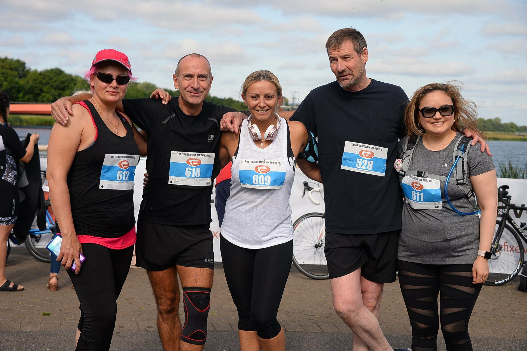 A group of five people, with race numbers 612, 610, 609, 528, and 611, pose for a photo outdoors. They are dressed in athletic wear, suggesting participation in a race or event. The background features a body of water and other participants.