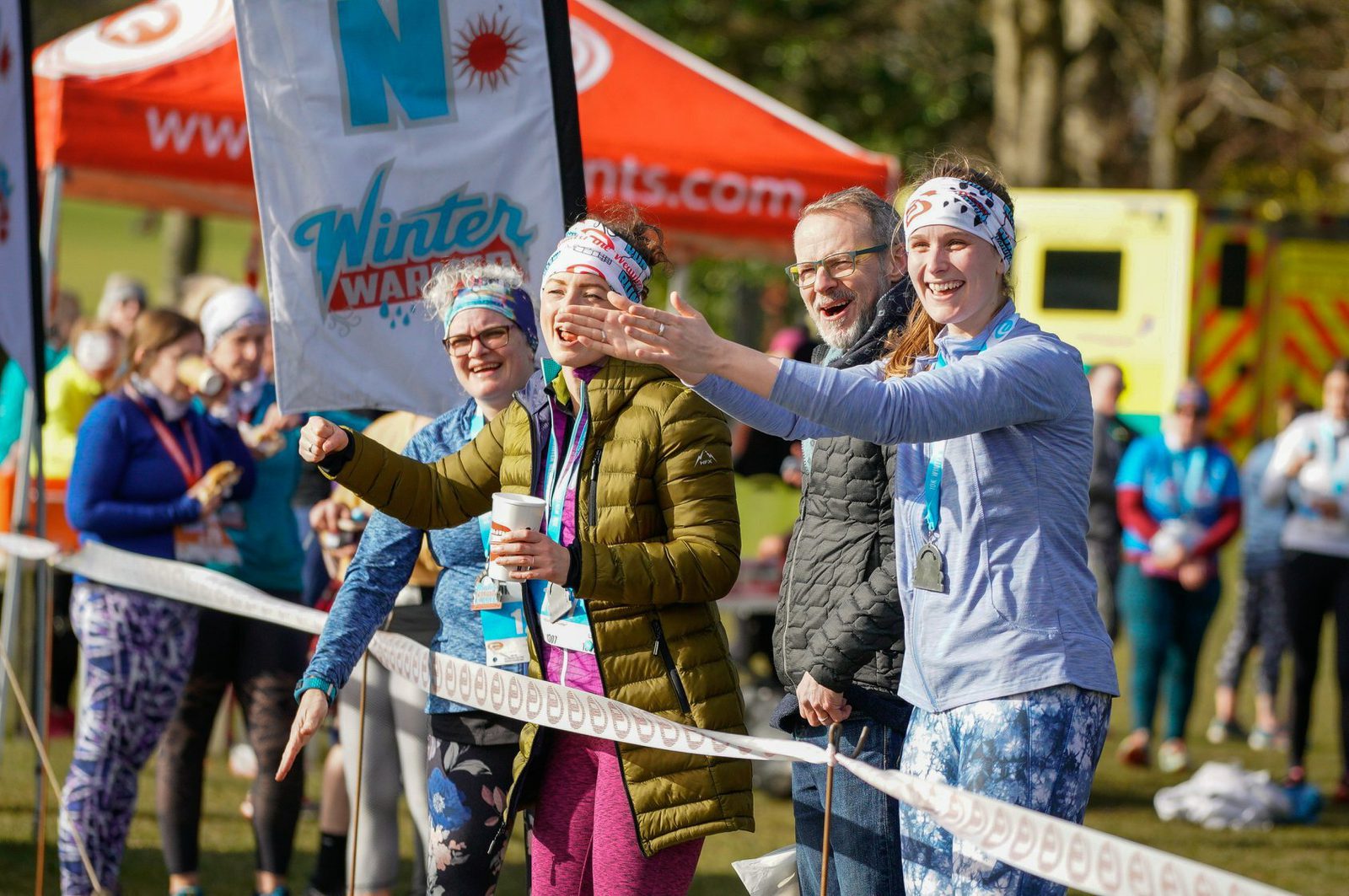 A group of jubilant runners celebrate near the finish line of an outdoor Winter Warmer event. They are dressed in athletic gear and wear medals around their necks. They stand behind a barrier with an orange tent and an emergency vehicle in the background.