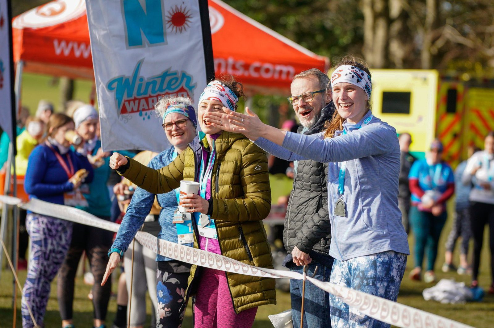 A group of enthusiastic people standing near a finish line, cheering and smiling during a winter run event. They are wearing medals, headbands, and athletic clothing. A red tent and other participants are visible in the background.