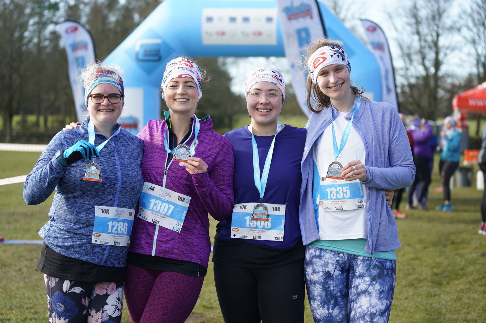 Four women in athletic wear pose together, smiling and holding medals at the finish line of a race event. They are wearing bib numbers, headbands, and medals around their necks. An inflatable arch and cheering spectators are visible in the background.