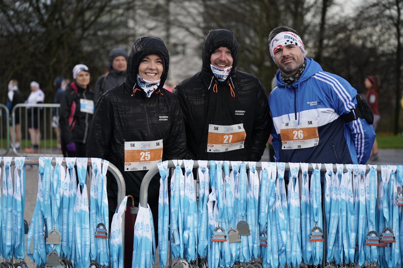 Three people wearing race bibs numbered 25, 26, and 27 are dressed in athletic gear and standing behind a rack filled with blue and white medals after a race. They are smiling and appear to be proud of their accomplishment. Trees and other participants are in the background.
