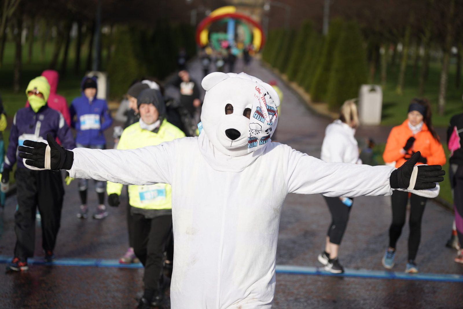 A person in a polar bear costume with arms outstretched is participating in a marathon. Other runners dressed in vibrant athletic wear are seen in the background, running on a paved, tree-lined path. An inflatable archway can be seen in the distance.