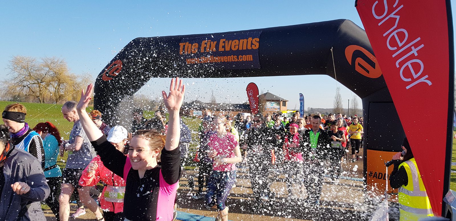 Participants in a fun run celebrate as they pass under an inflatable arch from "The Fix Events" labeled "www.thefixevents.com". Some runners have their hands raised and are running through a foam spray. A Shelter charity banner is visible on the right.