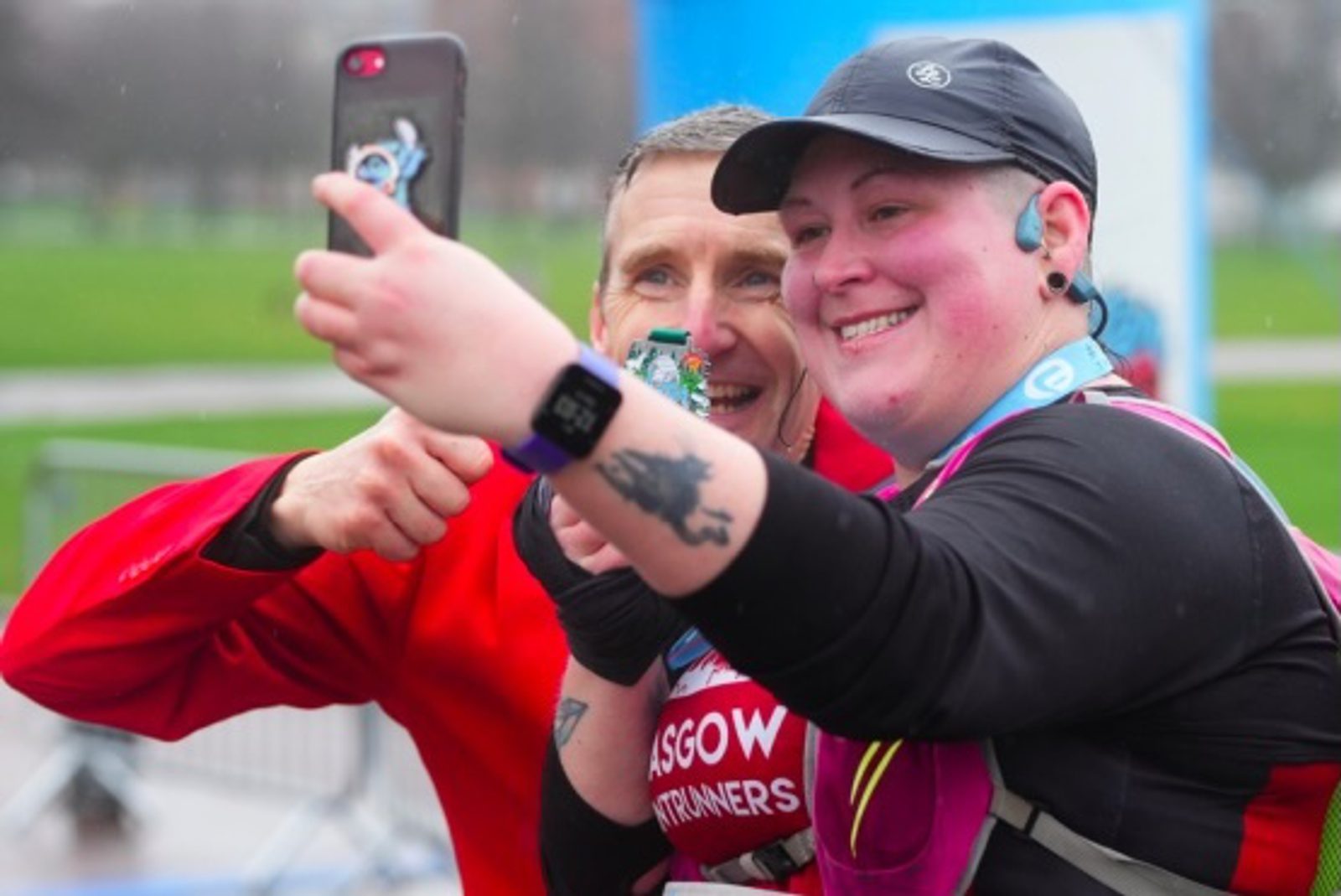 Two runners smile and take a selfie together after a race, one holding a medal. Both appear happy and are wearing running gear. The background is outdoors, slightly blurred, suggesting a park or race event area.