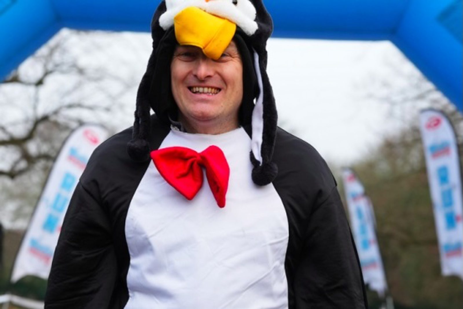 A person wearing a penguin costume with a red bow tie smiles while standing outdoors at an event with flags and an inflatable arch in the background.