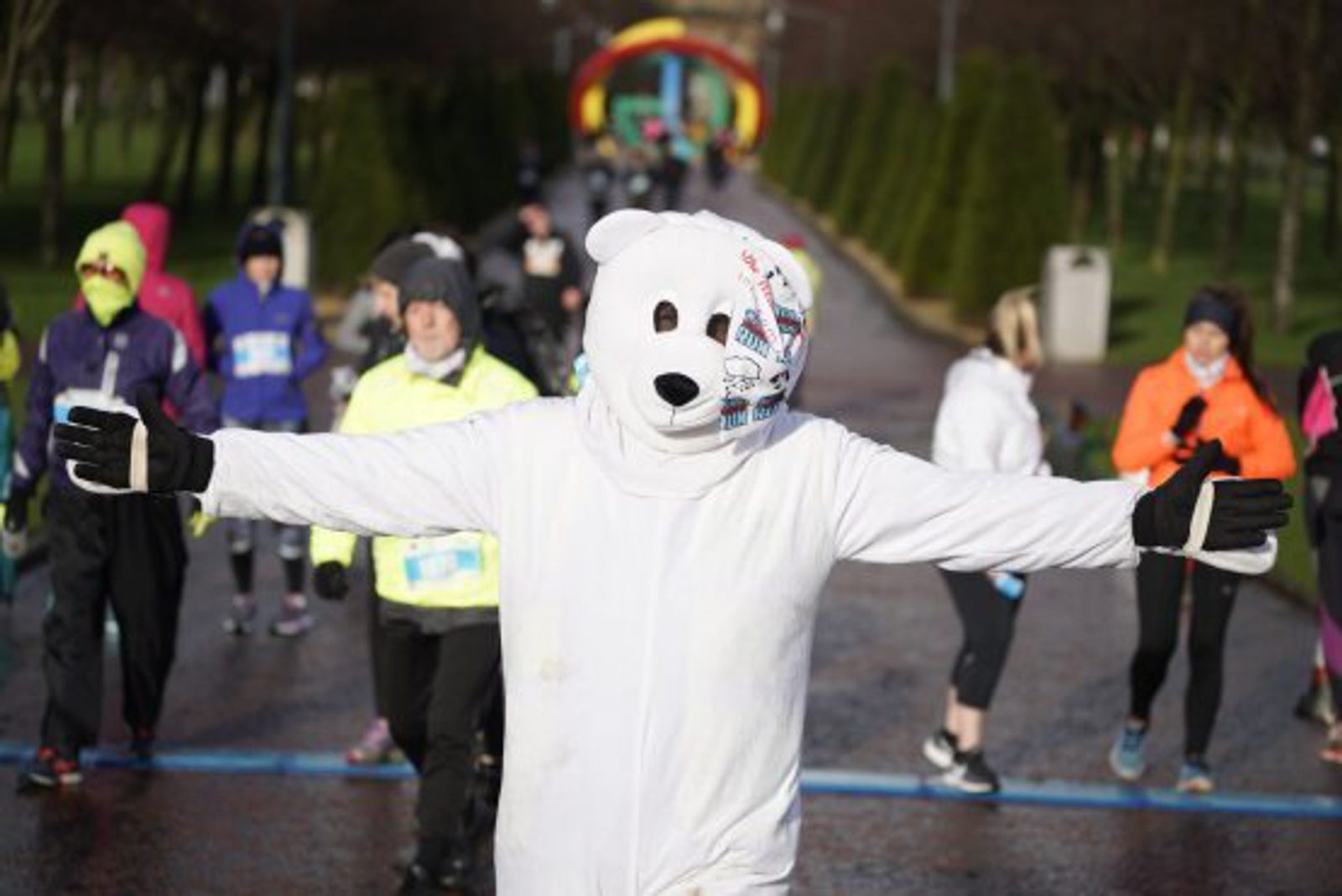 A person in a white polar bear costume stands with arms outstretched at a race event, while other runners in colorful athletic gear gather behind on a rainy road.