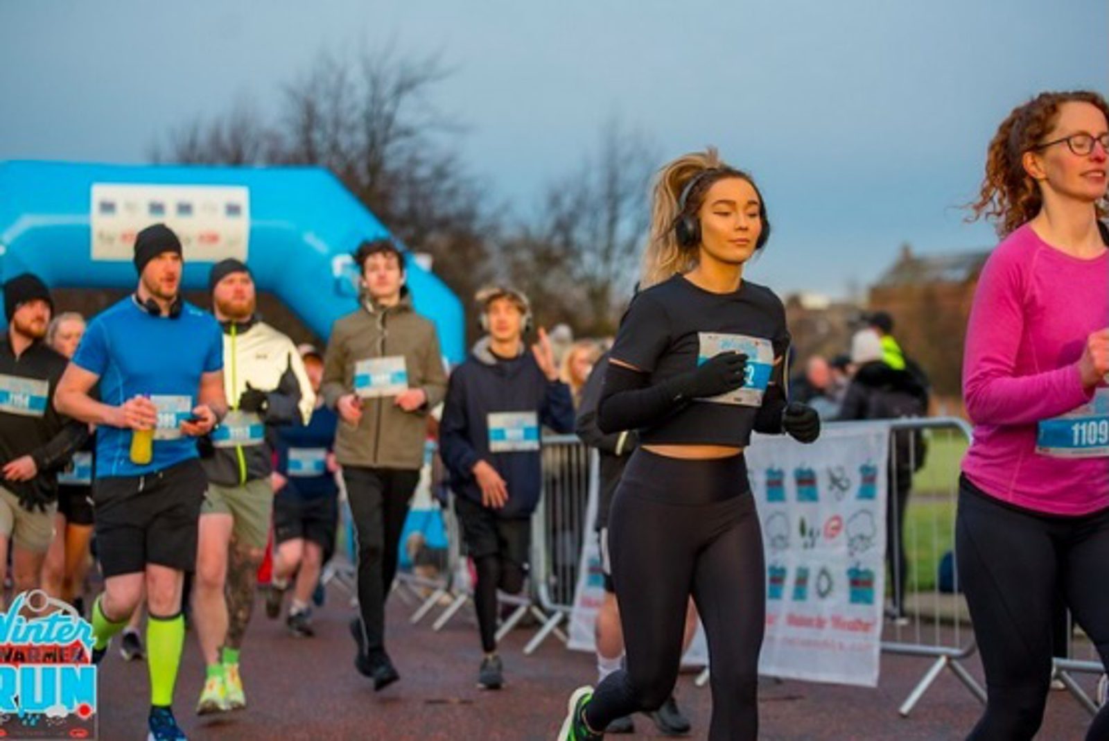 A group of runners, wearing race bibs and athletic gear, participate in an outdoor race with a blue inflatable archway in the background. Some runners wear headphones and it appears to be a cool, cloudy day.