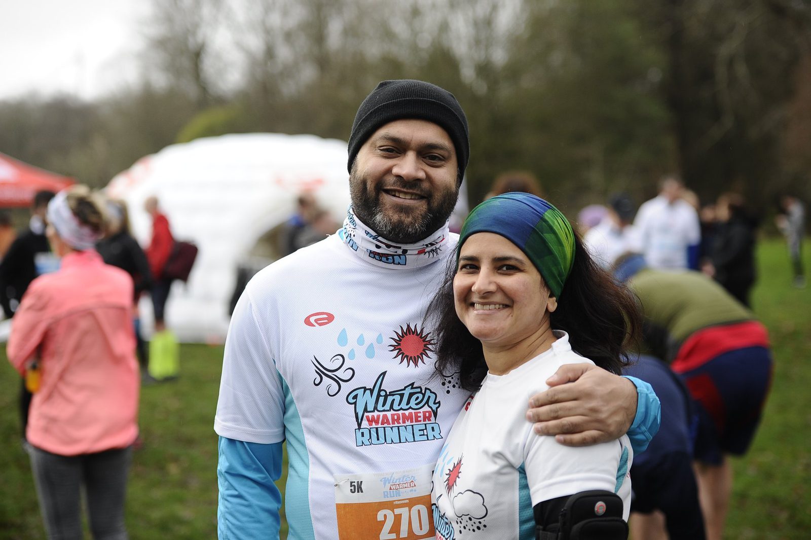 A smiling man and woman, both wearing white "Winter Warmer Runner" shirts and headbands, pose together outdoors at a running event. The man has his arm around the woman's shoulder. Other people and event tents are visible in the background.