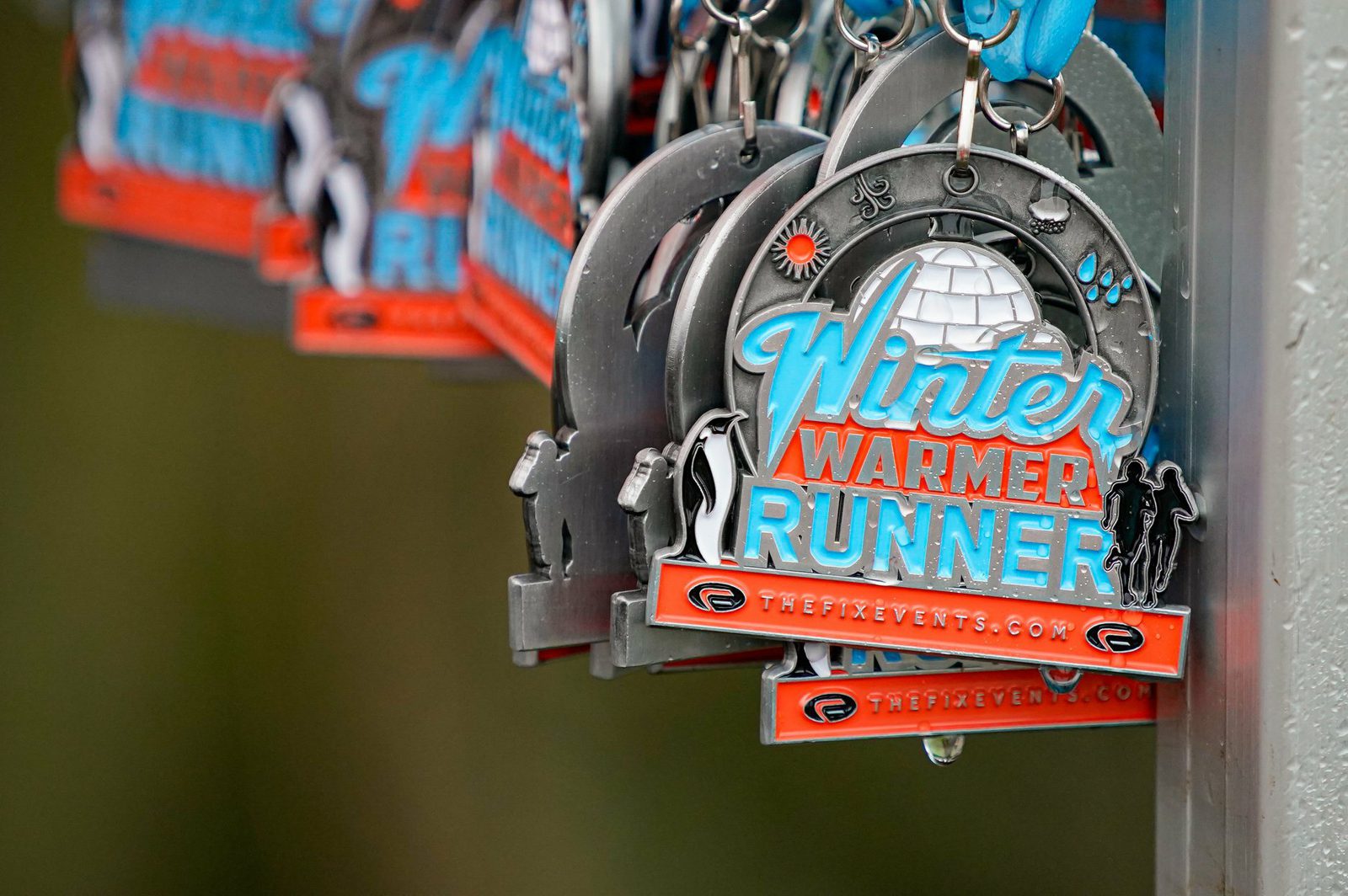 Close-up of several "Winter Warmer Runner" medals hanging. The medals feature a polar design with an igloo, snowflakes, and runners alongside the event logo and website. The medals are detailed with blue and orange colors and a ribbon for wearing.
