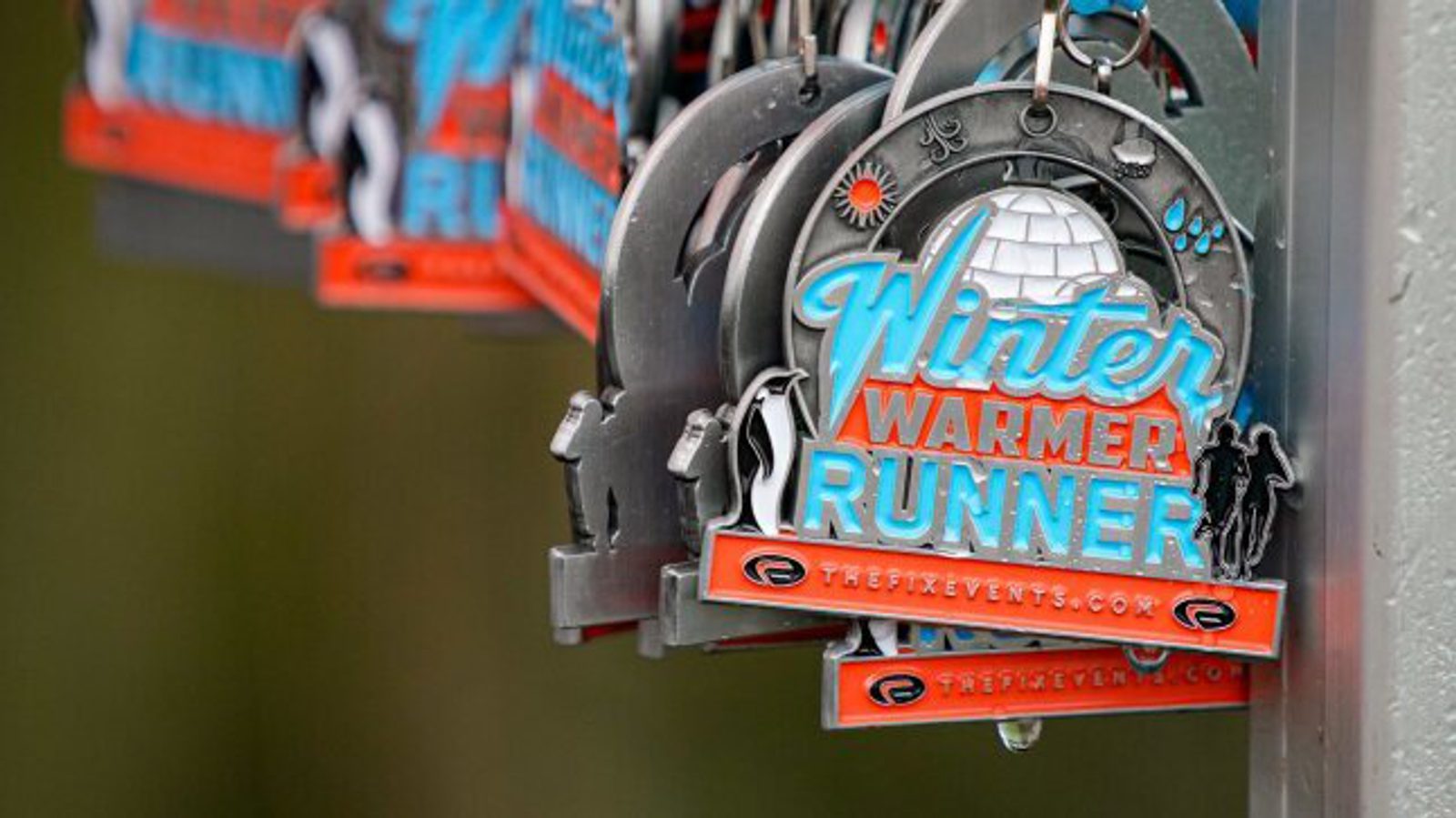 Multiple "Winter Warmer Runner" medals hang from hooks, featuring an igloo design with snowflakes, and the event website "thefitevents.com" prominently displayed. The medals have a winter theme with blue, orange, and grey colors.